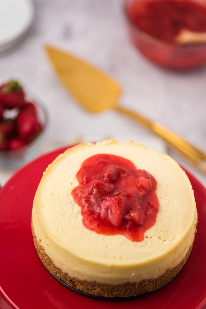 Instant pot cheesecake with strawberry jam topping on cake stand. strawberries and cake cutter in the background