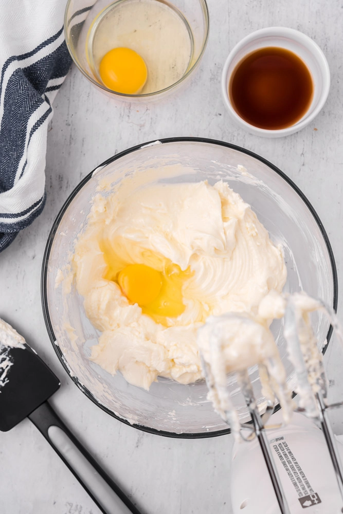 Cheesecake batter in bow with hand mixer. Egg in a bowl, vanilla extract ib a bowl, spatula.