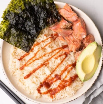 Rice and salmon bowl with seaweed, sriracha and avocado on a white plate with napkin and chopsticks