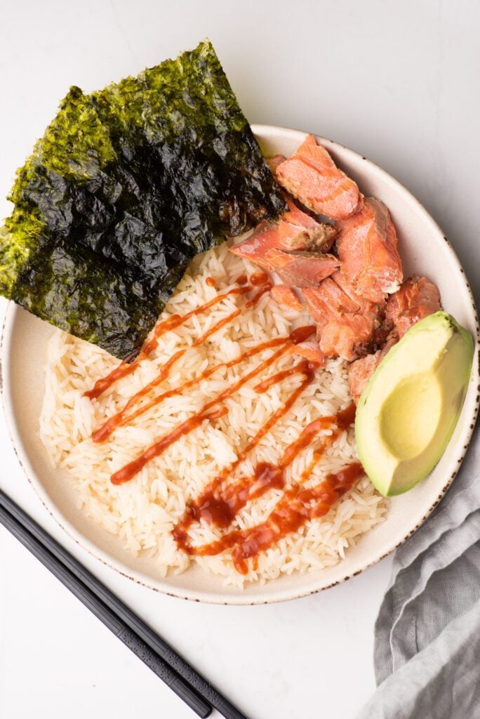 Rice and salmon bowl with seaweed, sriracha and avocado on a white plate with napkin and chopsticks