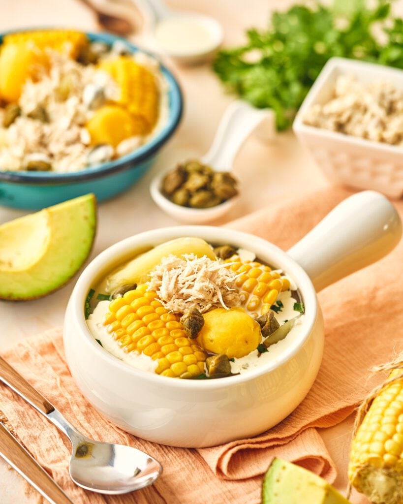 Colombian ajiaco with corn, chicken, potato and capers on a bowl with avocado, capers and bowl of ajiaco in the background