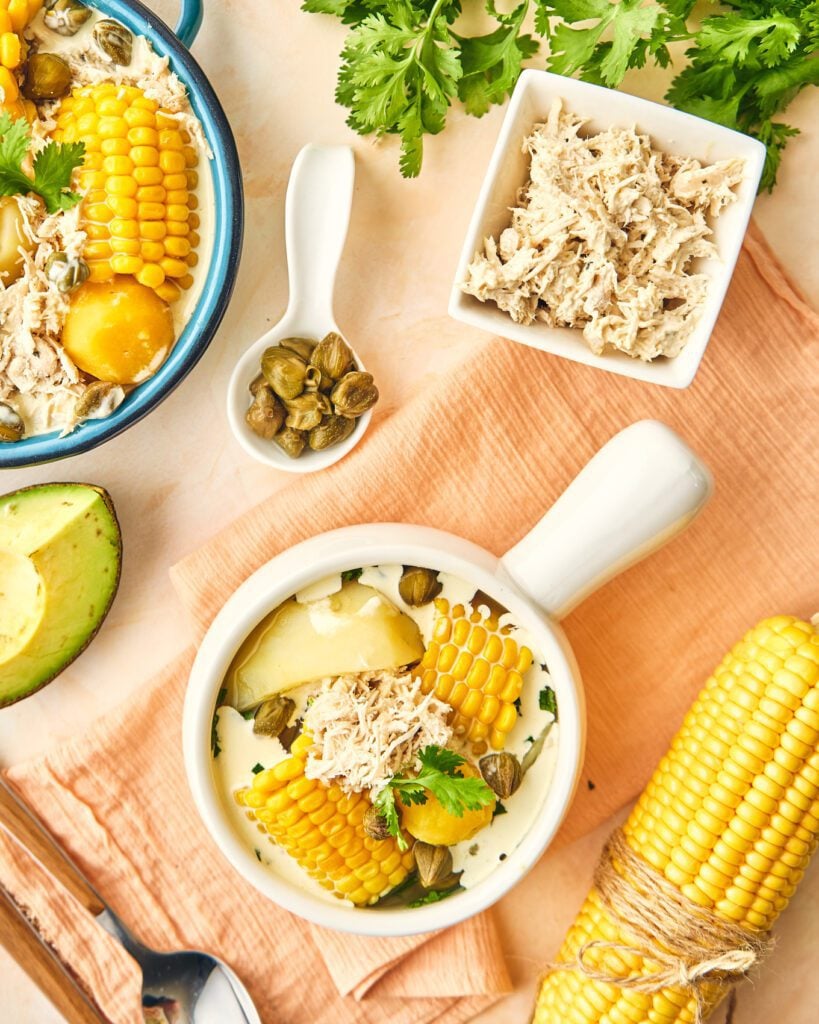 Ajiaco on a bowl with an ear of corn. Capers in a pinch bowl, shredded chicken in a bowl, sliced avocado