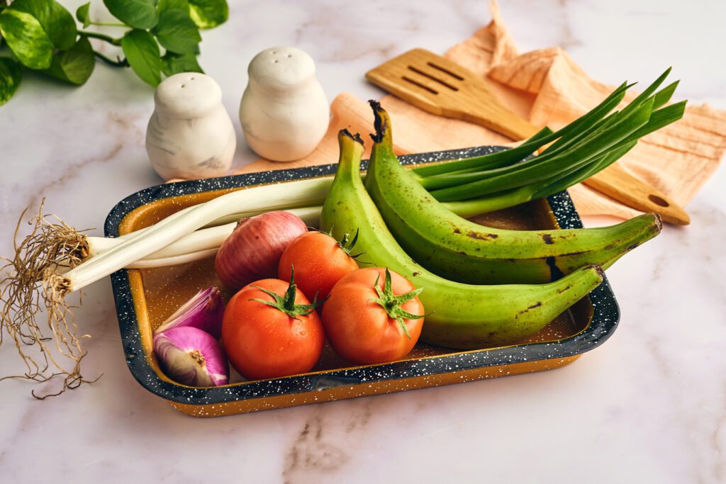 Bowl with onion, tomatoes, scallions and plantains. salt and pepper and wooden spoon on the background