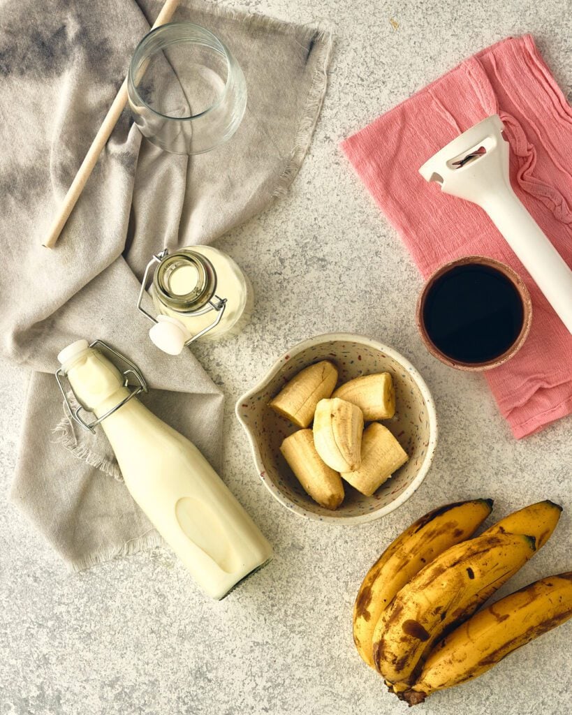 Bananas in a bowl, unpeeled bananas, oat milk and maple syrup. Blender, glass, straw and napkin