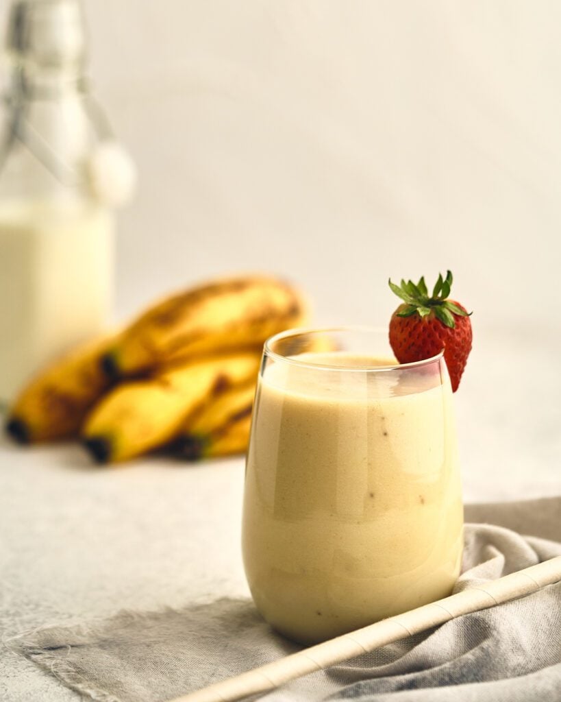 banana drink in glass with straw on the side. Strawberry on glass rim. Bananas on the back