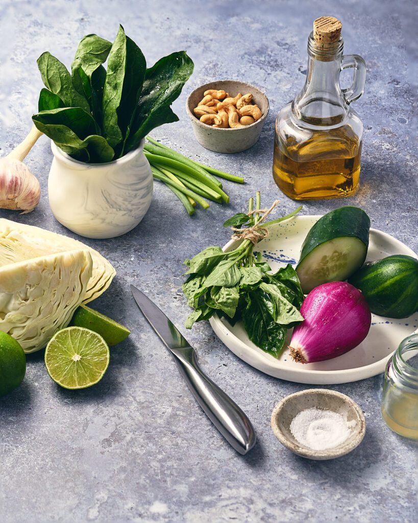 plate with shallots, cucumber and spinach next to knife and pinch bowl with salt with wedges of lime, vinegar in a glass, cashews in a bowl and cabbage wedge