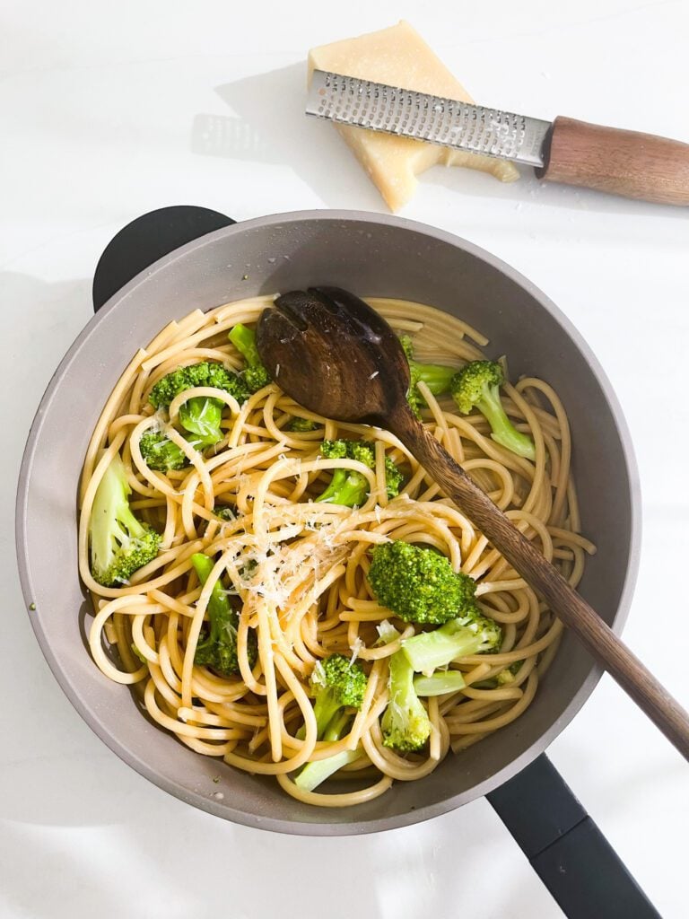 overhead view of broccoli with spaghetti in a skillet with wooden spoon. Parmesan with grater on the side