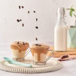 Whipped coffee with milk and spoon over a cutting board with coffee beans falling from above
