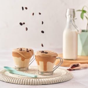 Whipped coffee with milk and spoon over a cutting board with coffee beans falling from above