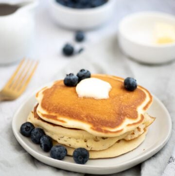 Pancakes on a plate with maple syrup, butter and blueberies placed on a white plate with napkin underneath