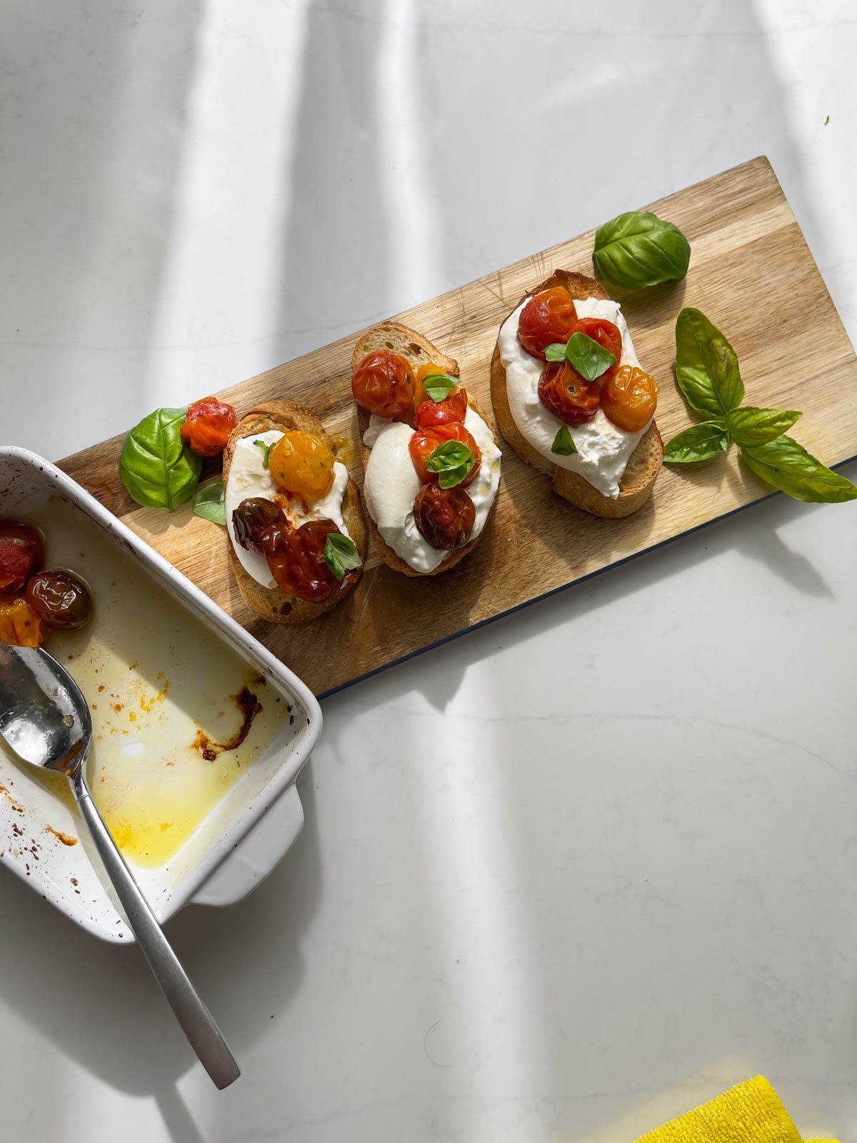 Jammy tomatoes on a baking dish next a wood board with bread topped with cheese, tomatoes and basil