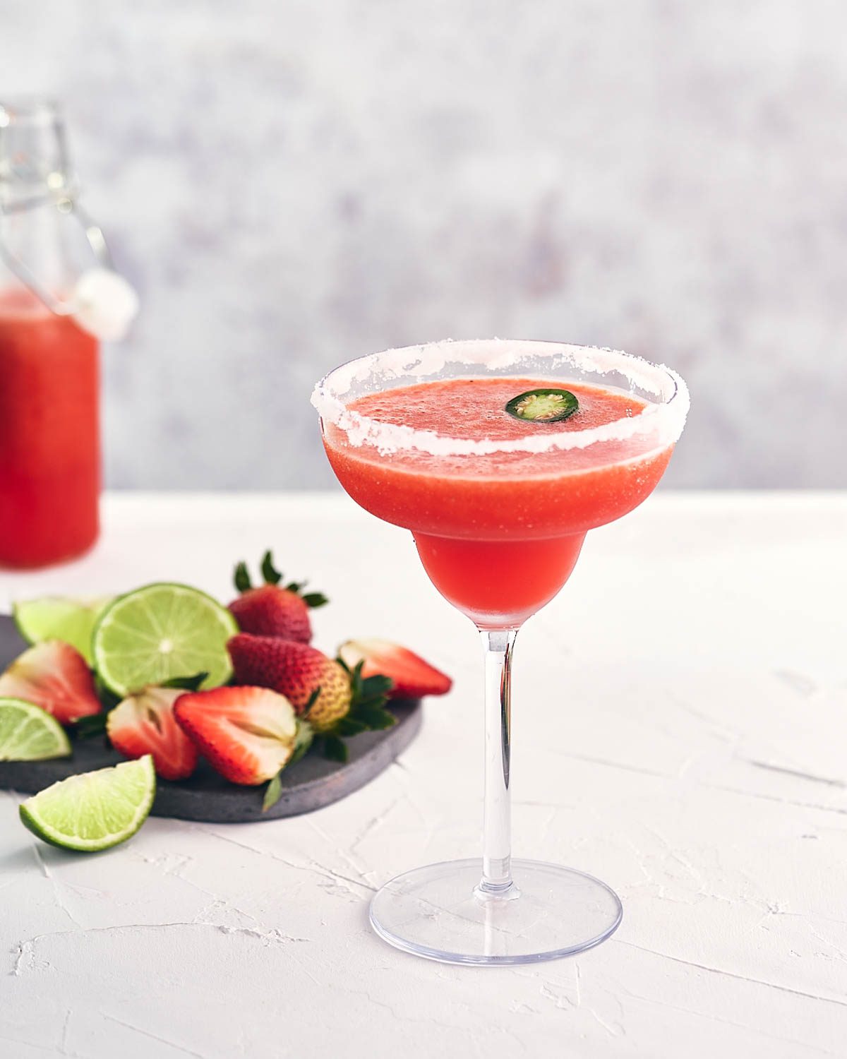 Margarita in a glass with jalapeño slice and limes and strawberries on a cutting board