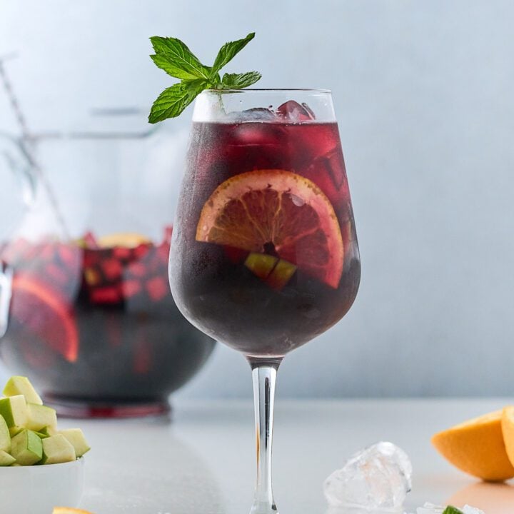 Sangria with orange slices and mint in wine glass with sangria pitcher in the back.