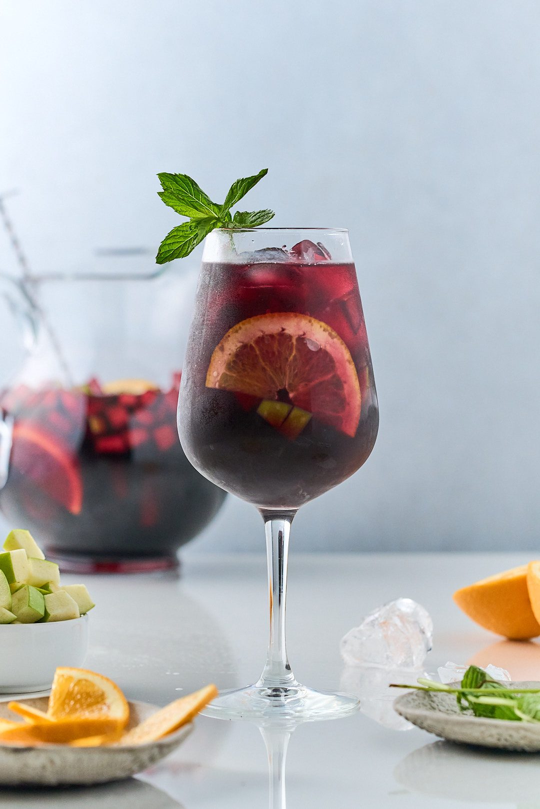 Sangria with orange slices and mint in wine glass with sangria pitcher in the back.