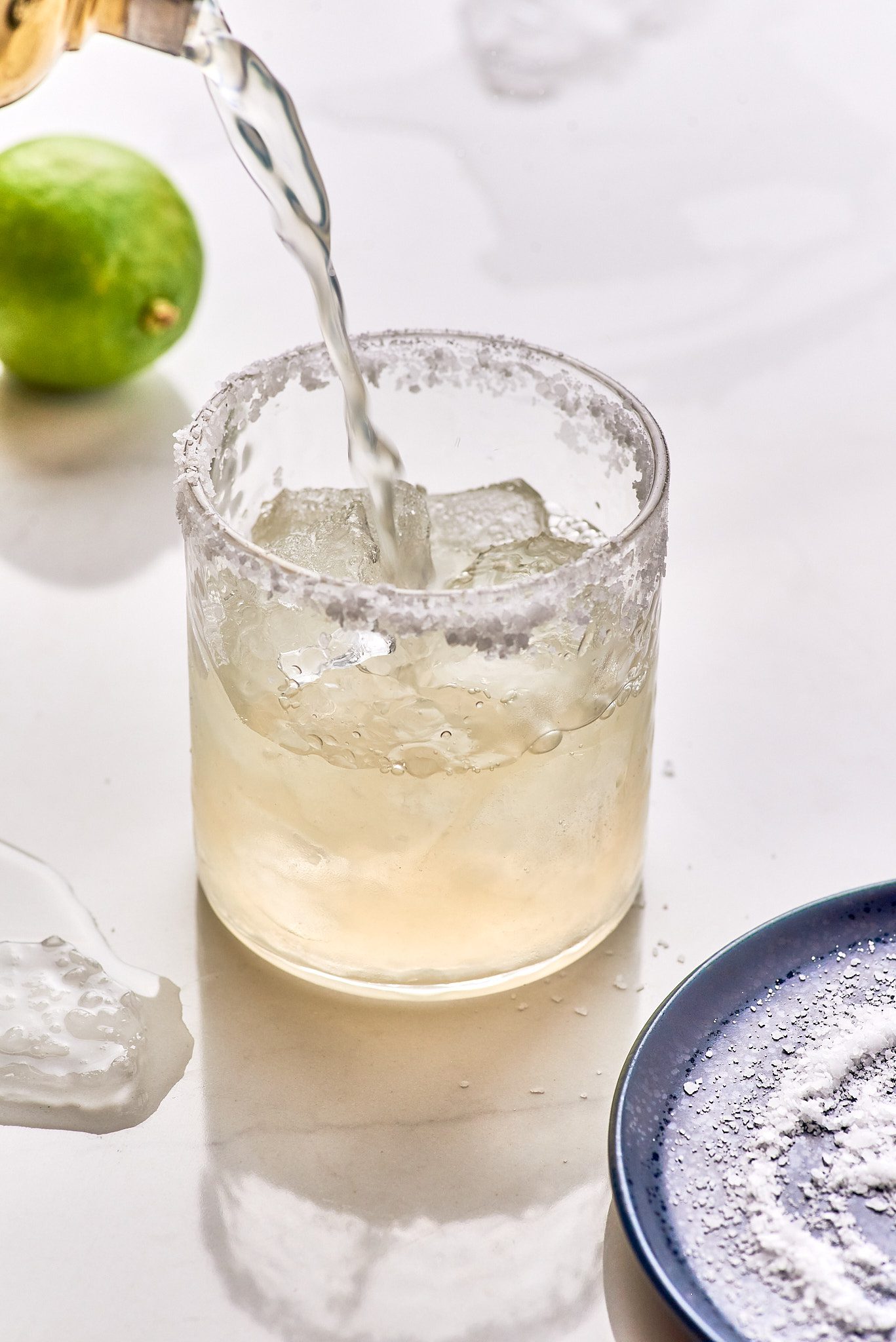 Margarita with salt rim on a glass. Lime on the background and plate with salt in the foreground.