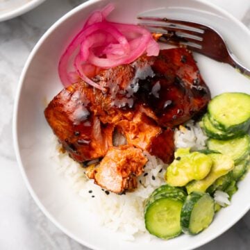 Teriyaki glazed salmon on a bowl served over rice with cucumbers and pickled onions.