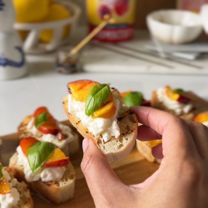 Peaches, burrata and bread with basil being held by hand with a wooden board in the background.