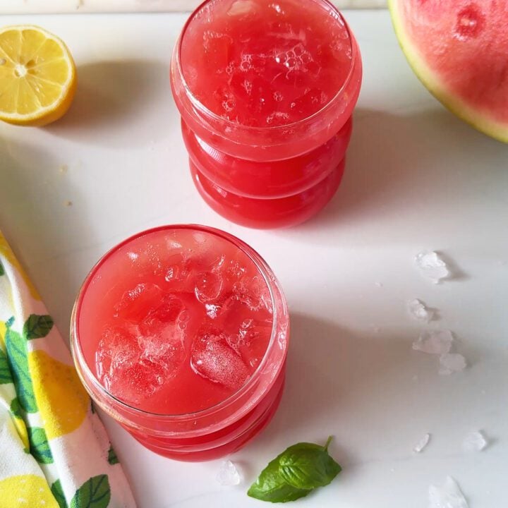 Watermelon lemonade on 2 glasses with lemon wedges and cut up watermelon in the background.