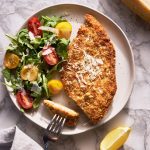 Crispy chicken cutlet in a plate with arugula and tomatoes garnished with parmesan cheese. Lemon slices as garnish and fork.