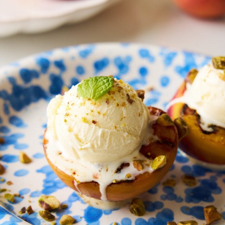 Grilled peaches with vanilla ice cream and chopped pistachios garnished with mint on a blue plate.